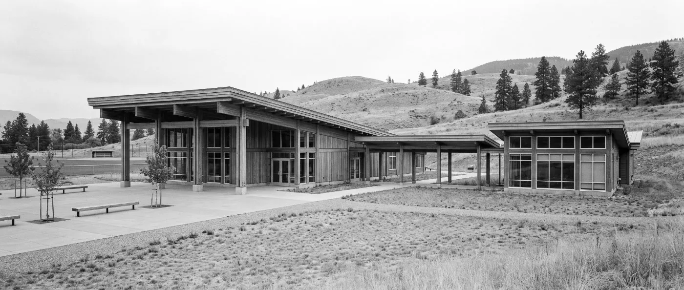 A modern First Nations community campus: a community hall with a broad cedar-clad roof on heavy timber posts, joined by a covered walkway to the administration building, grassland and plateau hills behind